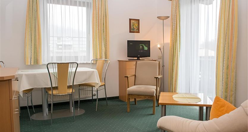 A bright living room with large windows and a cozy seating area. There is a table with chairs and a television.