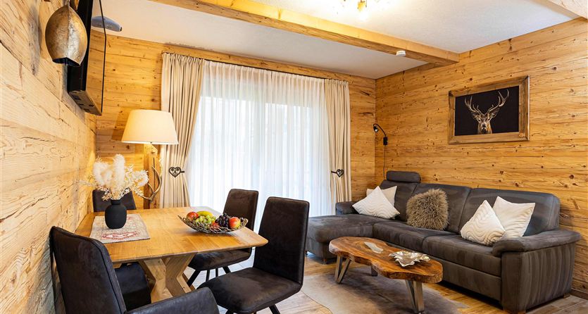 Ein gemütlicher Wohnraum mit Holzwänden, einem Sofa und einem Esstisch. Große Fenster sorgen für viel Licht und eine einladende Atmosphäre.