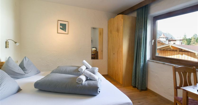A bright room with a double bed and two pillows. The room features a wardrobe and a window with a view outside.