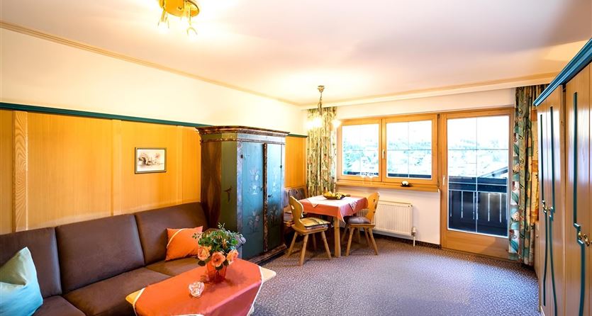 Ein gemütlicher Wohnraum mit einem Sofa, Holzmöbeln und einem kleinen Tisch. Große Fenster bieten einen Blick nach draußen und lassen viel Licht herein.