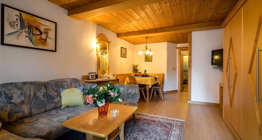 Ein gemütliches Wohnzimmer mit einem Sofa, einem Holztisch und einem Essbereich. Die warmen Farben und die Holzdecke schaffen eine einladende Atmosphäre.