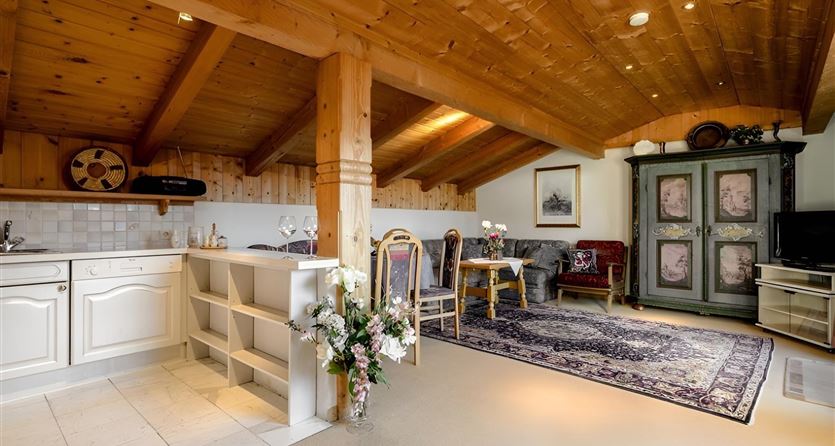 Ein gemütliches Zimmer mit Holzdecken und einem offenen Wohnbereich. Die Einrichtung umfasst eine Küchenzeile, einen Esstisch und einen alten Schrank.
