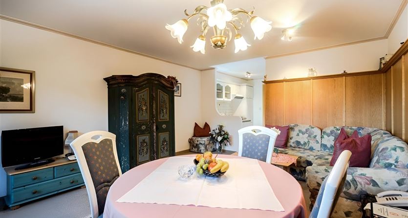 Ein gemütliches Wohnzimmer mit einem Esstisch, Stühlen und einer Couch. Der Raum ist hell beleuchtet und verfügt über eine antike Kommode sowie einen Fernseher.