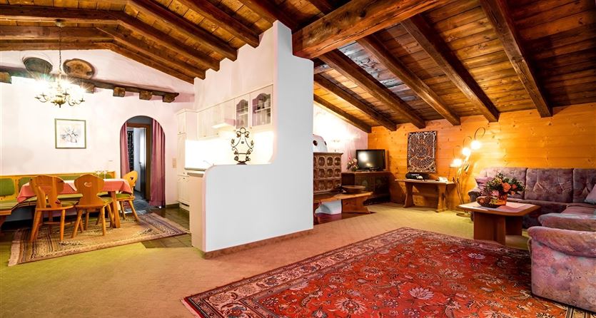 Ein gemütliches Wohnzimmer mit Holzbalkendecke und einem großen Teppich. Im Hintergrund befindet sich eine Esszeile mit einem Tisch und Stühlen.