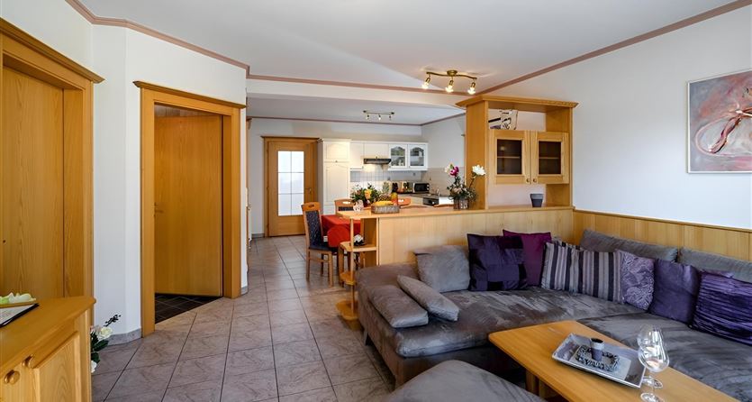 Ein gemütliches Wohnzimmer mit einem großen Sofa und einem Esstisch. Die Raumgestaltung ist hell und modern, mit Holzdetails und einer offenen Küche.