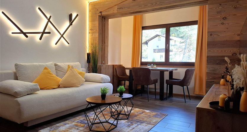 A modern living room with a comfortable sofa, cool color tones, and warm light. A dining table and a window with a view of nature create an inviting atmosphere.