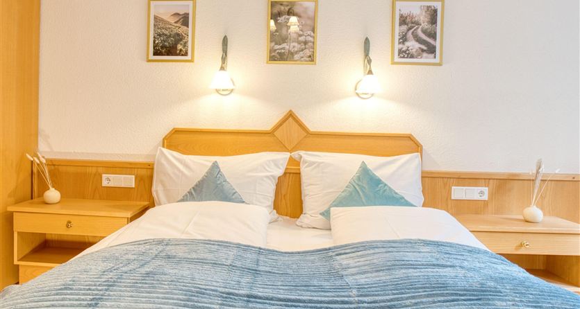 A cozy bedroom with a double bed and blue pillows. Pictures hang on the walls, and there are nightstands with lamps.
