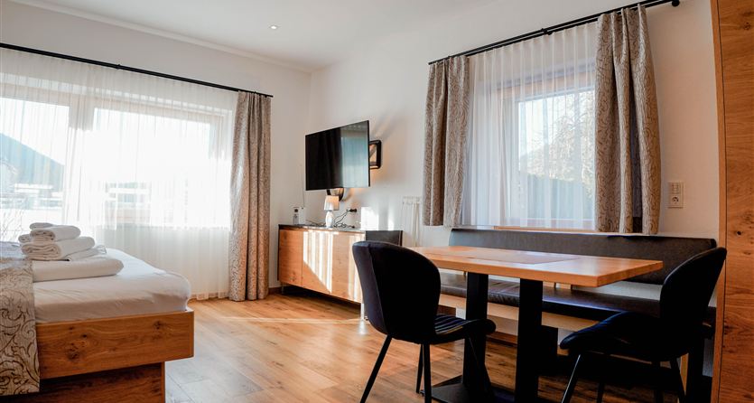 Ein modernes Hotelzimmer mit einem großen Bett, einem Tisch und Stühlen. Große Fenster sorgen für viel Tageslicht.