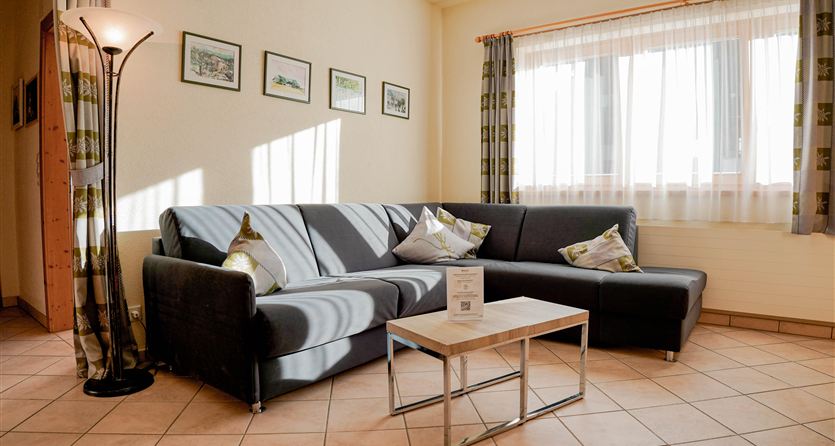 Ein gemütliches Wohnzimmer mit einem grauen Sofa und einem Glastisch. Sanftes Licht strömt durch die Fenster, umgeben von dekorativen Vorhängen.