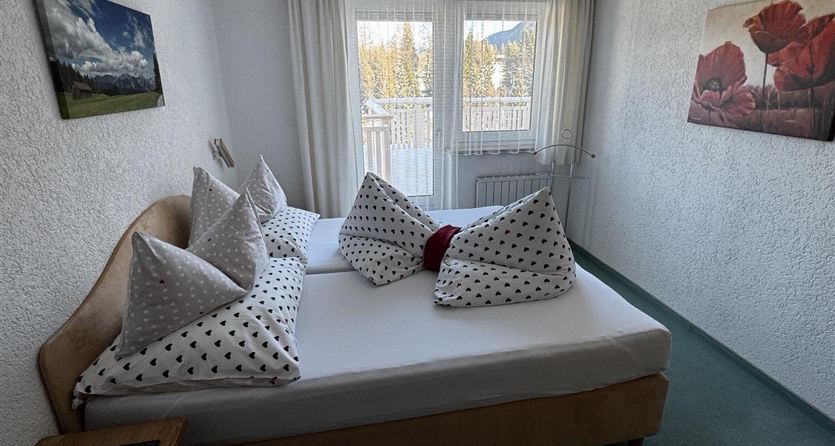 Ein gemütliches Schlafzimmer mit zwei Betten, dekorierten Kissen und einem großen Fenster. An den Wänden hängen Bilder mit Landschaften und Blumen.