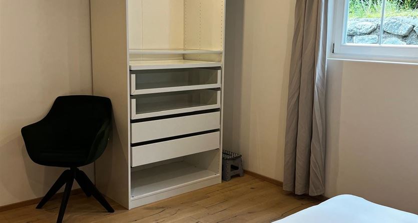 A modern bedroom with a minimalist wardrobe and a comfortable chair. The wooden floor and the curtains create a cozy atmosphere.