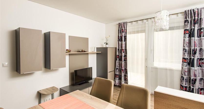 Een moderne woonkamer met een eetgedeelte en een televisie. Grote ramen zorgen voor veel daglicht en de gordijnen geven de ruimte een frisse uitstraling.