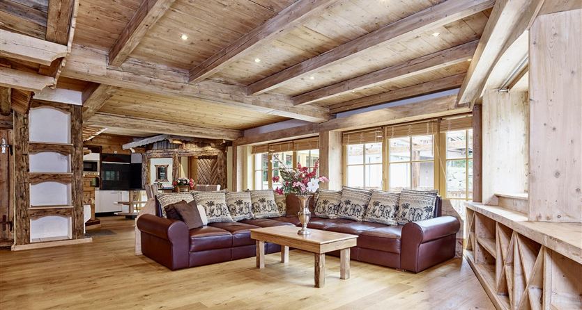 Een gezellige woonkamer met houten balken en een groot raam. Een donkere bank met kussens en een houten tafel maken de ruimte compleet.
