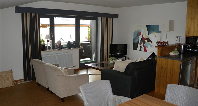 A modern living room with a black sofa and a dining table. Large windows let in plenty of light and provide a view outside.