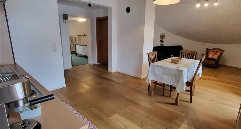 Un luminoso e spazioso soggiorno con pavimento in legno e un tavolo per quattro persone. Sullo sfondo si vedono mobili e una cucina open space.