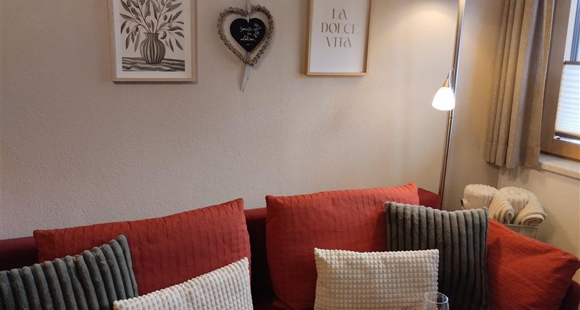 Een gezellig woonkamer met een rode bank en verschillende decoratieve kussens. Aan de muur hangen schilderijen en een hartvormige lijst.
