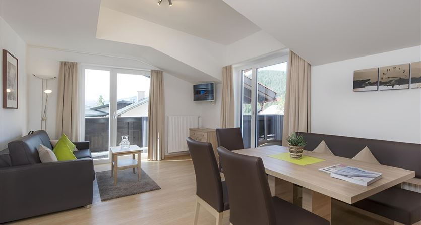 Eine moderne Wohnküche mit einem gemütlichen Sofa und einem Esstisch. Große Fenster sorgen für viel Licht und einen Blick nach draußen.