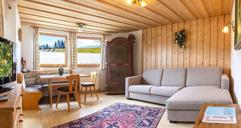 Ein gemütliches Wohnzimmer mit Holzvertäfelung, einem bequemen Sofa und einem Esstisch. Große Fenster bieten einen Blick ins Grüne.