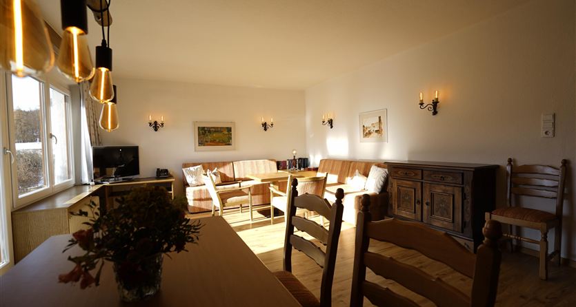 Een heldere woonkamer met comfortabele meubels en een groot raam. Op de tafel staat een bloemenvaas en de muren zijn versierd met lichtbronnen en schilderijen.