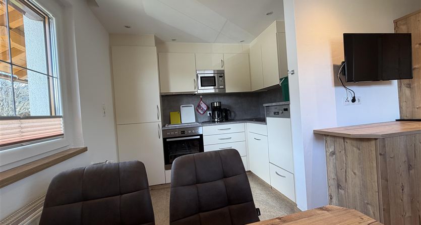 Een moderne keuken met witte kasten en apparaten. Op de voorgrond staan twee stoelen aan een houten tafel.
