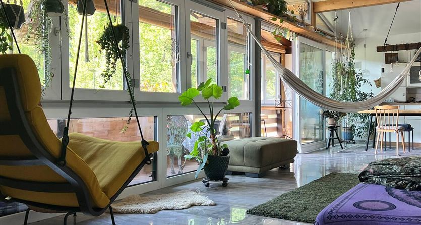 A cozy room with large windows, plants, and a hammock. There is a modern armchair and a soft carpet on the floor.