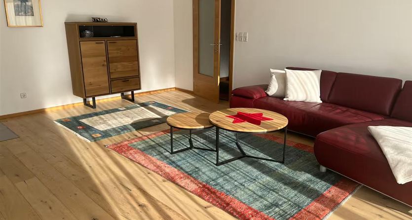 Ein helles Wohnzimmer mit einem roten Sofa und einem Holzregal. Auf dem Boden liegt ein Teppich mit einem bunten Muster und zwei kleinen Tischen.