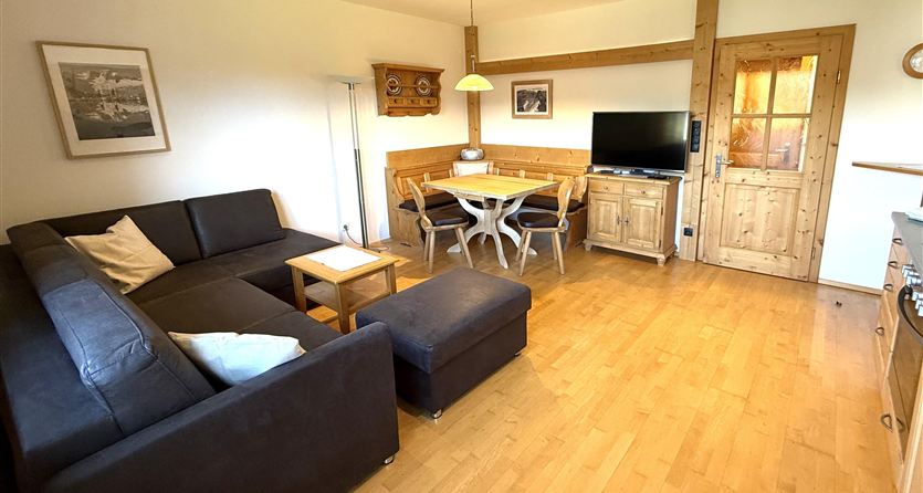 Ein gemütlicher Wohnbereich mit einem schwarzen Sofa und einem Holztisch. An der Wand steht ein Fernseher, und das Licht fällt durch ein Fenster.