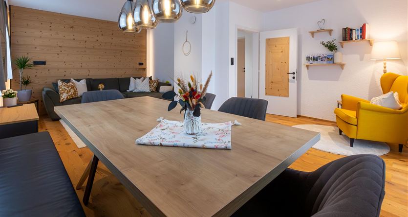 Een modern woonkamer met een grote houten tafel en comfortabele stoelen. Een gele stoel, planten en decoratieve elementen geven de ruimte een gezellige sfeer.