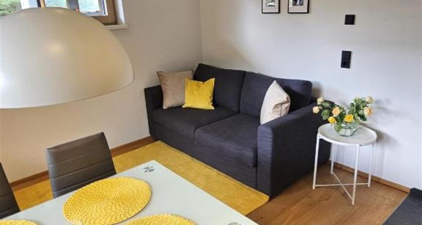 Ein gemütlicher Wohnbereich mit einem grauen Sofa und gelben Kissen. Ein Tisch mit gelben Platzsets und einer eleganten Lampe schafft eine freundliche Atmosphäre.