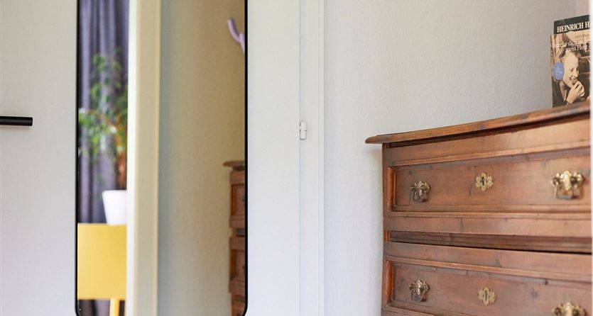 Ein heller Raum mit einer weißen Tür, die ein rechteckiges Fenster hat. Neben der Tür steht eine alte Kommode aus Holz.