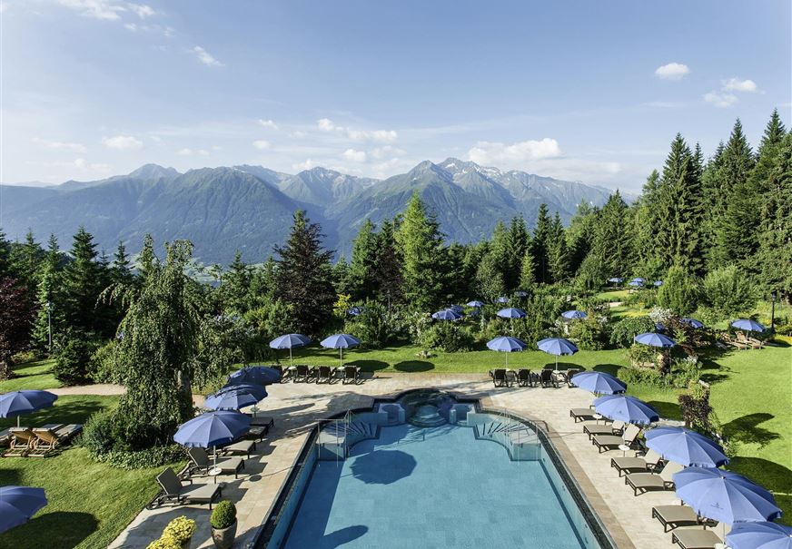 A beautiful pool surrounded by a well-kept garden and mountains in the background. Sun umbrellas and loungers invite you to relax.
