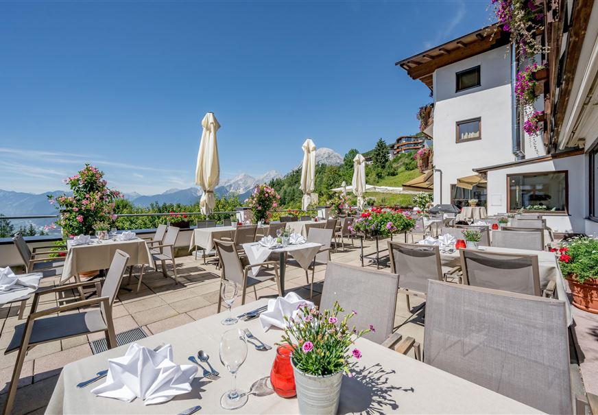 Ein einladendes Restaurant mit vielen Tischen im Freien und bunten Blumen. Im Hintergrund sind Berge und ein klarer blauer Himmel zu sehen.