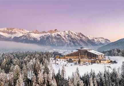 A modern hotel in a snow-covered landscape with majestic mountains in the background. The sky is clear and has a soft hue at sunrise.