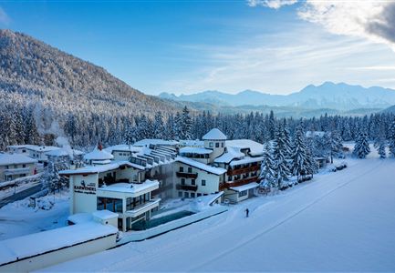 Eine idyllische winterliche Landschaft mit schneebedeckten Bäumen und Bergen im Hintergrund. Im Vordergrund sind gemütliche Gebäude zu sehen, die in der ruhigen Umgebung liegen.