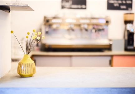 A yellow vase with dried flowers stands on a modern counter. In the background, a coffee machine and a cozy café atmosphere are visible.