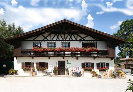A traditional Bavarian house with a balcony full of flowers. The facade is white and brown, surrounded by green nature.