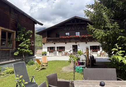 A traditional house in the Alps with a large garden and terraced furniture. In the foreground are chairs and tables for relaxing outdoors.